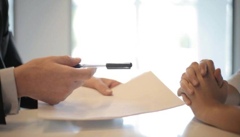 A person in a suit handing over a document and pen to another person who is clasping their hands.